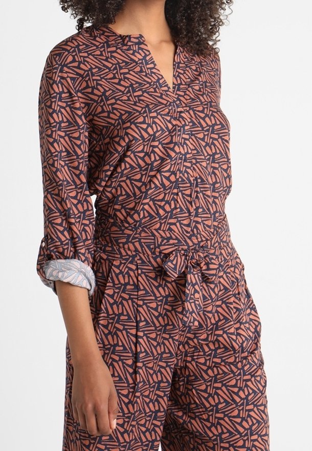 Tuta alla moda con maniche lunghe e colletto, caratterizzata da un motivo geometrico nei colori rosso, blu navy e crema; il materiale è leggero, con una vita regolata da una cintura e dettagli con risvolto sulle maniche.