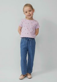Meisje staat rechtop en draagt een lavendelkleurig, gebloemd shirt met korte mouwen, blauwe jeans met elastische taille en beige ballerina's tegen een effen achtergrond.
