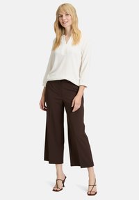 Chemisier blanc texturé à manches longues avec un col en V. Pantalon large marron. Modèle portant des sandales noires à brides. Fond neutre.
