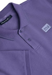 Lila polotröja med texturerad tyg, två knappar, en stickad krage och ett fyrkantigt logomärke på bröstet.