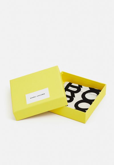 Kollane ristkülikukujuline kast valge kleebisega, millel on kiri "MARC JACOBS." Kasti sees on osaliselt nähtav must-valge mustriga ese.