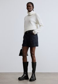 Bel bel prstan pulover z visokim ovratnikom, temno modre kratke jeans hlače in črni škornji do kolen z debelimi podplati. Majhna črna torbica v levi roki.