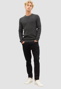 Donkergrijs gebreid sweater met een ronde halslijn, gecombineerd met zwarte broeken en witte sneakers, staande tegen een grijze achtergrond.