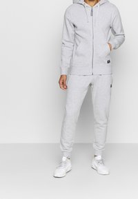 Hellgrau zip-up Kapuzenpullover mit Kordelzügen und Fronttaschen, kombiniert mit passenden schmal geschnittenen Sweatpants und weißen Sneakers. Glatte Textur.