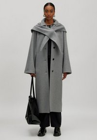 Grauester Wollmantel mit breitem Kragen und übergroßer Silhouette, ausgestattet mit großen schwarzen Knöpfen und einem strukturierten Design. Kombiniert mit einer schwarzen Tasche.