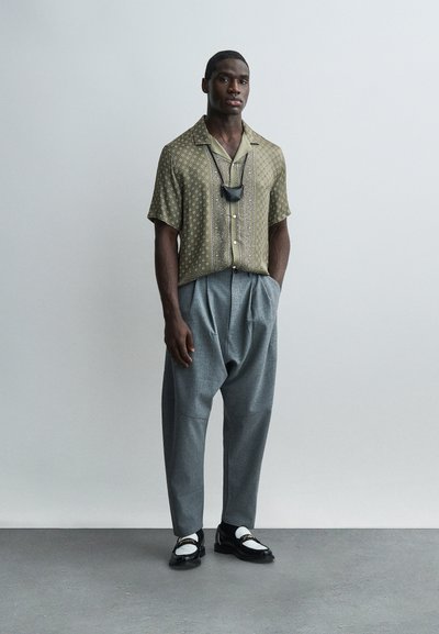 Άνδρας που στέκεται μπροστά σε γκρίζο τοίχο, φοράει πουκάμισο με σχέδιο και κοντά μανίκια, γκρι πλισέ παντελόνια, μαύρα και λευκά μοκασίνια και μαύρη μάσκα προσώπου κρεμασμένη γύρω από τον λαιμό.