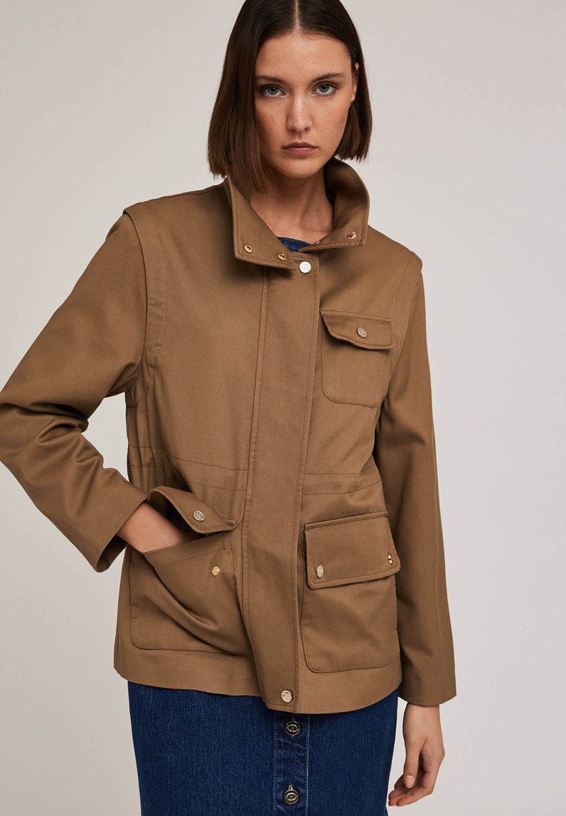 Brązowa bawełniana kurtka z wysokim kołnierzem, zatrzaskami i dwiema przednimi kieszeniami; gładka faktura z wyraźnie zarysowanym krojem.