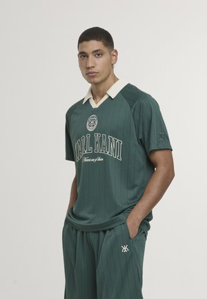 Joven con camisa verde oscuro de rayas finas de Karl Kani y pantalones a juego, de pie con las manos en los bolsillos frente a un fondo blanco.