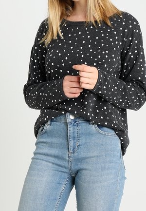 Person trägt einen dunkelgrauen Pullover mit weißen Punkten und hellblaue, hochtaillierte Jeans, die Hände vor der Taille verschränkt vor einem schlichten Hintergrund.