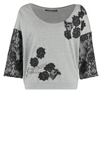 Haut femme gris clair avec motifs floraux en dentelle noire sur les manches et le devant, manches trois-quarts et large encolure ronde.