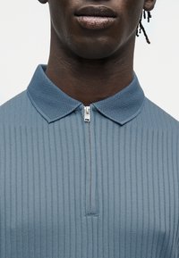 Κοντινό πλάνο ενός ατόμου που φοράει μπλε ριμπ φιλέ κοντομάνικο με φερμουάρ και υφή στο γιακά, πάνω σε απλό φόντο.