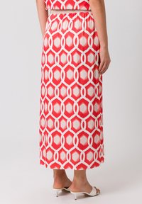 La jupe maxi présente un motif géométrique rouge et blanc, avec une texture lisse et une taille élastique. Elle est associée à des sandales à talons blanches.