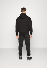 Felpa nera con cappuccio e pantaloni cargo abbinati. Il materiale è una miscela di cotone. Dettagli notevoli includono tasche laterali e sneaker con motivo.