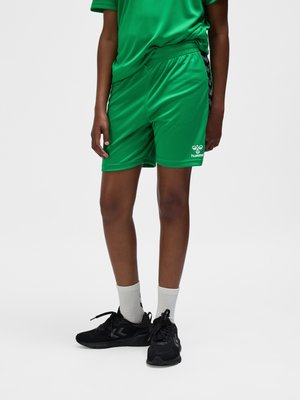 Person, der bærer grønne sportsshorts og matchende skjorte, hvide sokker og sorte sportssko, stående på en ensfarvet lys baggrund.