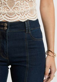 Jeans a vita alta blu scuro con cuciture dorate a contrasto, caratterizzati da due bottoni decorativi, abbinati a un top in pizzo color crema.