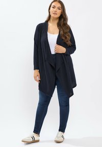 Cardigan de canalé en color navy con apertura frontal, mangas largas y un diseño fluido. Combinado con unos jeans oscuros ajustados y unas zapatillas casuales beige con rayas negras.