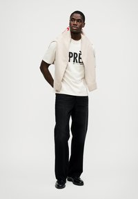 Λευκό T-shirt με στάμπα "PRÈ", μπεζ πουλόβερ περασμένο γύρω από τους ώμους, μαύρο τζιν και μαύρα παπούτσια. Απλός σχεδιασμός και ουδέτερη χρωματική παλέτα.