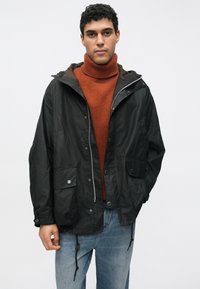 Schwarze wasserdichte Jacke mit Kapuze, Druckknöpfen, Tunnelzug an der Taille und zwei Vordertaschen. Über einem rostorangenen Rollkragenpullover getragen.