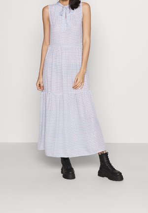 Frau trägt ein ärmelloses weißes Maxikleid mit kleinem blauem Muster und schwarzen klobigen Schnürstiefeln, steht vor einem schlichten Hintergrund.
