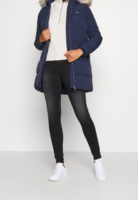 Marinblå puffjacka med pälsbeklädd huva, dragkedjeslutning och framfickor. Krämfärgad stickad tröja, mörka jeans och vita sneakers fulländar outfiten.