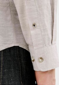 Lys beige skjorte med et struktureret stof. Indeholder manchetter med to knaplukninger og sort syning. Sort denimmateriale, der delvist er synligt.
