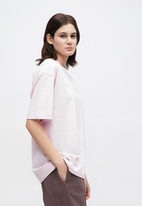 Νέα γυναίκα με καστανά μαλλιά μέχρι τους ώμους, φορώντας ένα χαλαρό ροζ μπλουζάκι και καφέ παντελόνι, στέκεται με το ένα χέρι στην τσέπη μπροστά σε λευκό φόντο.