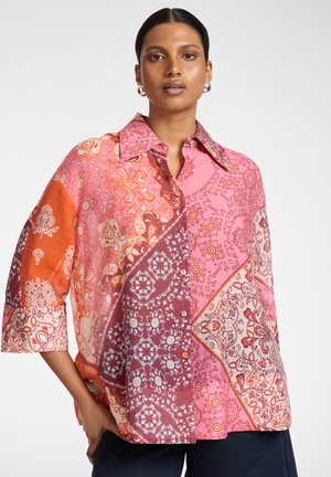 Donna con capelli scuri raccolti indietro che indossa orecchini di perle e una camicia ampia multicolore in patchwork con motivi rosa, arancioni e bordeaux.