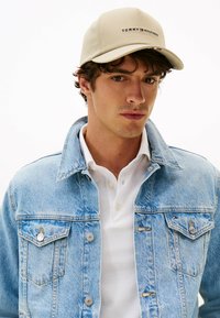 Young man wearing beige Tommy Hilfiger cap, light blue denim jacket, and white polo shirt, facing forward against a plain background.