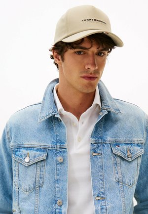 Young man wearing beige Tommy Hilfiger cap, light blue denim jacket, and white polo shirt, facing forward against a plain background.