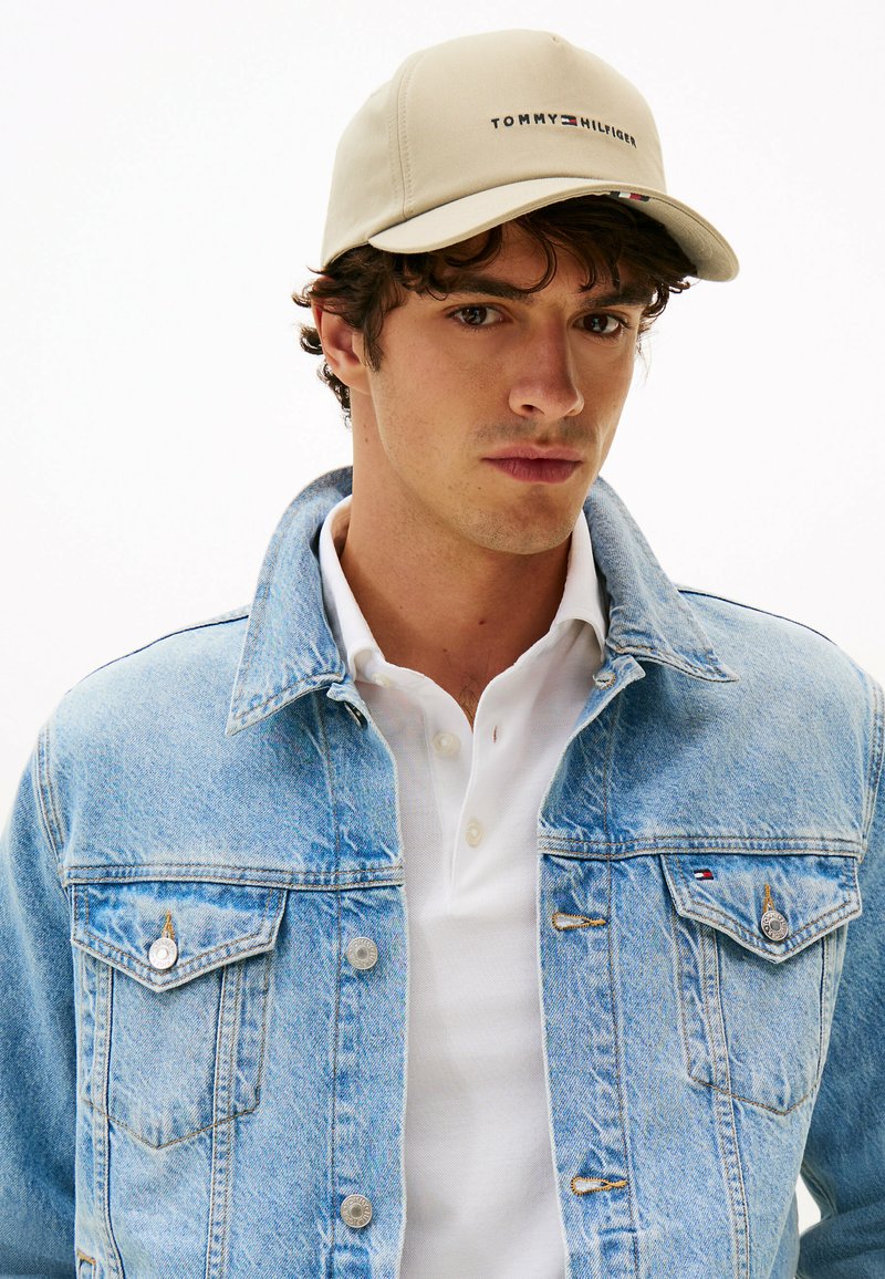 Young man wearing beige Tommy Hilfiger cap, light blue denim jacket, and white polo shirt, facing forward against a plain background.