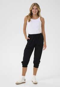 Černé joggingové kalhoty s elastickými manžetami a páskem, s detailem na šňůrku, kombinované s bílým tílkem a bílými sportovními botami.