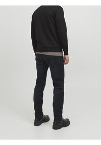 Uomo in piedi di spalle che indossa una felpa nera, jeans blu scuro, una maglietta marrone sotto e scarpe di pelle nere, su sfondo neutro.