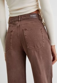 Ruskeat farkuhousut, joissa on korkea vyötärö ja suora lahje. Housuissa on kaksi takataskua ja brändätty tarra vyötäröresorissa.