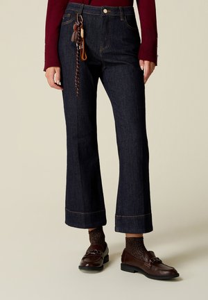 Jeans dritti in denim scuro con orli risvoltati, indossati con mocassini marroni, calzini con texture e una maglia a maniche lunghe color bordeaux con polsini abbottonati.