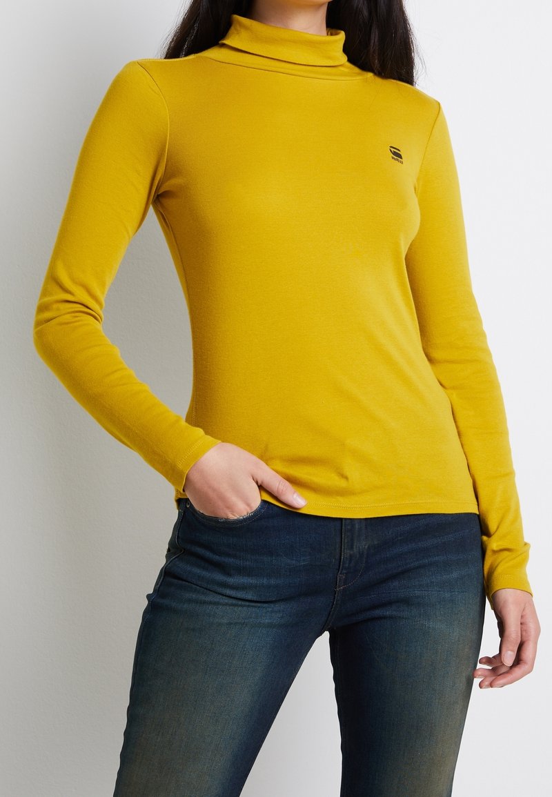 Gelbes Langarm-Turtleneck-Oberteil aus glattem Stoff, mit taillierter Passform und kleinem Logo auf der Brust. Kombiniert mit dunklen Jeans.