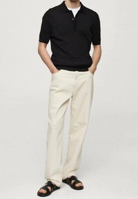Czarna prążkowana koszulka polo z krótkim rękawem, zestawiona z beżowymi szerokimi spodniami i czarnymi sandałami na paski, ręce w kieszeniach.