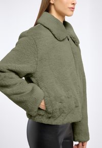 Przycięta, oliwkowa sztuczna futrzana kurtka z dużym kołnierzem i ukrytym zapięciem z przodu. Teksturowany materiał; widoczne boczne kieszenie.