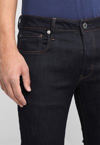 Jeans de mezclilla azul oscuro con un diseño ajustado, costuras naranjas y un cierre visible de botón y cremallera. Cuenta con bolsillos laterales y traseros.