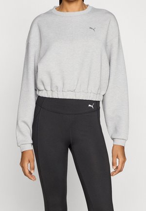 Kobieta nosi jasnoszarą krótką bluzę i czarne legginsy z wysokim stanem, oba z małymi logo Puma na piersi i biodrze.