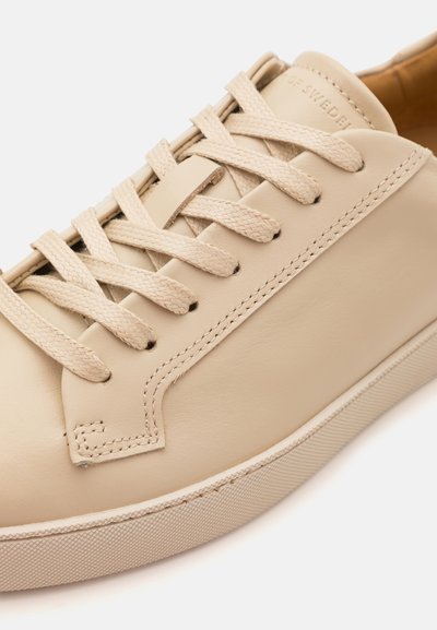 Beige leather sneaker with flat woven laces, smooth texture, and rubber sole. Features stitching detail along the sides and a minimalist design.