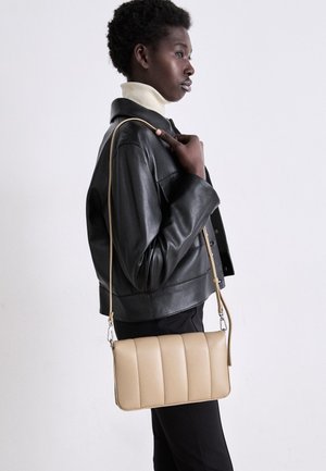 Person iført en sort læderjakke og en cremefarvet rullekrave sweater holder en beige quiltet skuldertaske med en lang rem.