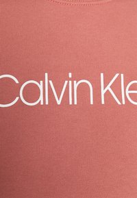 Logo w białym kolorze marki Calvin Klein, umieszczone na zbliżeniu różowego tła z tkaniny, prawdopodobnie odzieży.