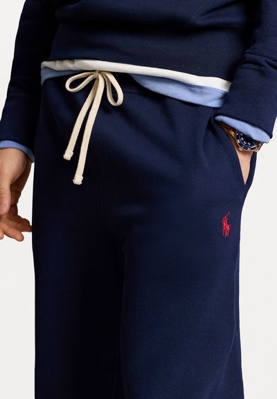 Pantalons de jogging bleu marine avec une taille élastique et un cordon de serrage crème. Présente un logo rouge brodé sur la cuisse supérieure et un tissu texturé.