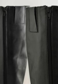 Schwarze Lederstiefeletten bis zum Knie verfügen über einen vollständigen Reißverschluss an der Außenseite und eine glatte Textur, mit kontrastierenden matten und glänzenden Oberflächen.