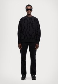 Felpa nera con design testurizzato, caratterizzata da un motivo ondulato. Abbinata a pantaloni neri e sneakers nere, indossata con occhiali da sole.