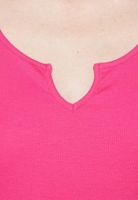 Dopasowany różowy top z prążkowanego materiału z płytkim dekoltem w kształcie litery V i kontrastowym przeszyciem. Tekstura jest gładka z subtelnymi pionowymi liniami.