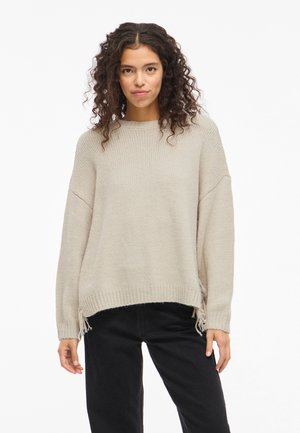 Beige knitted sweater with a relaxed fit, ribbed hem, and side slits featuring fringe details. Worn over black jeans.