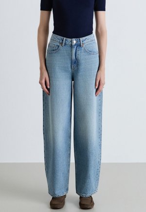 Persona che indossa jeans azzurri chiari a vita alta e a gamba larga abbinati a scarpe marroni e una maglietta scura a maniche corte su sfondo semplice.