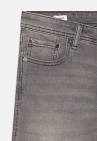 Jean en denim gris avec un design classique à cinq poches, des accents de boutons et une texture décolorée. Étiquette visible au niveau de la taille.