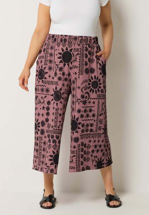 Frau trägt weite, knöchellange Hose in Mauve mit schwarzen Sonnen- und Blumenmotiven, kombiniert mit einem weißen T-Shirt und schwarzen Sandalen.